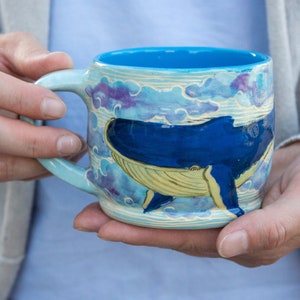 Puede incluir: Una taza de cerámica con interior azul y asa azul claro. El exterior presenta un diseño de ballena pintado a mano en tonos azules y amarillos, sobre un fondo de nubes estilizadas en azul y morado.