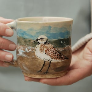 May include: A ceramic mug with a hand-painted shorebird design. The mug has a beige background with a coastal scene in blue, brown, and white. The bird is depicted with brown and white feathers. The mug is held in two hands.
