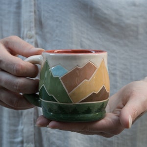 May include: A ceramic mug with a mountain scene design. The mug is painted with green, brown, yellow, and orange mountains, and a blue sky. The mug has a green base with a row of trees.