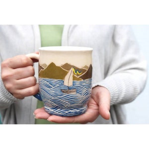 May include: A ceramic mug featuring a blue and white wave pattern with a sailboat on a sea against a backdrop of brown mountains.