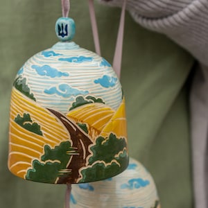 Puede incluir: Una campana de cerámica pintada a mano con una escena de paisaje de colinas onduladas, un camino sinuoso y un cielo azul con nubes. La campana está decorada con un símbolo de tridente ucraniano en la parte superior.