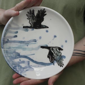 以下が含まれることがあります： 2 羽の黒い鳥が飛んでいる手描きのデザインが施された白いセラミックプレート。プレートには青い筆致と飛沫があり、ダイナミックで芸術的な美しさを生み出しています。プレートは人が持っています。