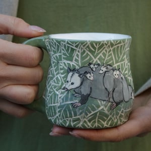 Puede incluir: Una taza de cerámica verde con interior blanco presenta un diseño pintado a mano de una zarigüeya madre llevando a sus crías. Las zarigüeyas están representadas en tonos de gris y blanco sobre un fondo verde de hojas.