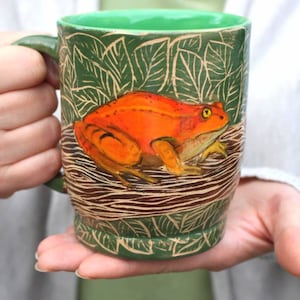 Könnte beinhalten: Grüne Keramiktasse mit Froschdesign. Die Tasse zeigt eine leuchtend orangefarbene Froschillustration mit gelben Augen, vor einem Hintergrund aus grünen Blättern und braunen, holzähnlichen Linien. Der Innenraum und der Griff sind ebenfalls grün.