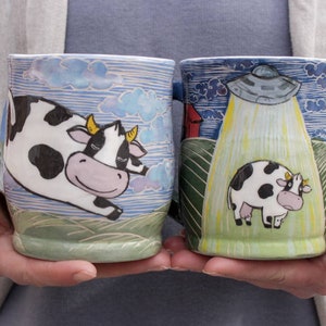 Puede incluir: Dos tazas de cerámica con un diseño caprichoso. Una taza presenta una vaca en blanco y negro volando por el aire con un cielo azul y nubes en el fondo. La otra taza presenta una vaca en blanco y negro de pie en un campo con un OVNI flotando sobre ella.