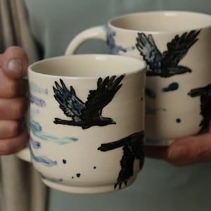 Könnte beinhalten: Zwei weiße Keramikbecher mit schwarzen Vogelmotiven und blauen Akzenten. Die Becher haben eine zylindrische Form mit Henkeln. Die Vogelillustrationen zeigen fliegende Vögel. Die Becher werden in den Händen einer Person gehalten.