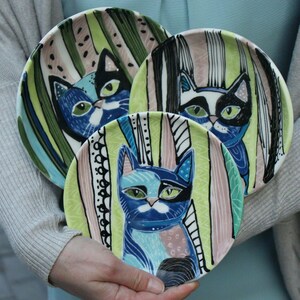 Könnte beinhalten: Drei Keramikplatten mit Katzenmotiven. Jede Platte zeigt ein anderes Katzen-Gesicht mit blauen, schwarzen und weißen Markierungen, vor einem Hintergrund aus grünen, rosa und weißen vertikalen Streifen. Die Teller werden vor einem hellblauen Hintergrund gehalten.