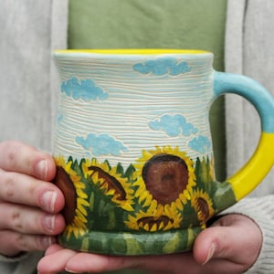 May include: A ceramic mug with a yellow rim and handle. The mug is painted with a blue sky, white clouds, and a field of sunflowers. The handle is painted yellow and blue.