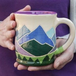 Puede incluir: Una taza de cerámica con un diseño pintado a mano de una cordillera en tonos azules, verdes y morados. La taza tiene un fondo blanco y un asa verde.