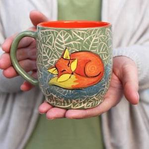 Peut inclure: Une tasse en céramique verte avec un bord orange et un motif peint à la main d'un renard endormi sur un lit de feuilles.