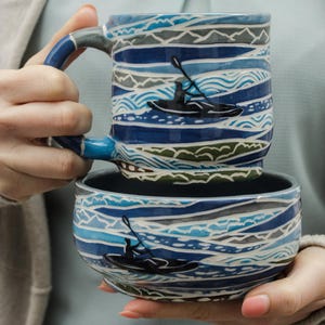 Puede incluir: Un juego de taza y tazón de cerámica azul y blanco con un patrón de olas y una silueta de una persona haciendo kayak. La taza y el tazón están apilados uno encima del otro.