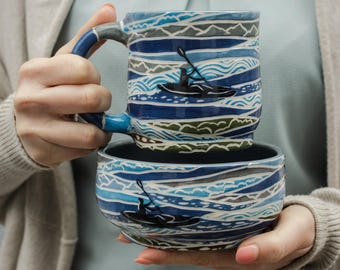 May include: A blue and white ceramic mug and bowl set with a wave pattern and a silhouette of a person kayaking. The mug and bowl are stacked on top of each other.