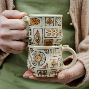 Puede incluir: Dos tazas de cerámica con fondo blanco y patrones geométricos naranja, marrón y negro. Las tazas están apiladas una encima de la otra.