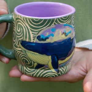 May include: A green ceramic mug with a swirling pattern and a painted image of a whale swimming in a blue and yellow ocean. The mug has a lavender interior.