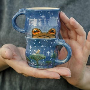 Peut inclure: Deux tasses bleues en céramique empilées, chacune avec une anse. Les tasses sont décorées d'étoiles blanches et d'un motif de grenouille. Les yeux de la grenouille sont orange. La tasse du bas a un motif de nénuphar.
