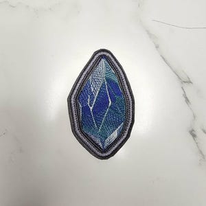 May include: A diamond-shaped embroidered patch featuring a faceted crystal design in shades of deep blue, teal, and white. The patch is outlined with a thick, dark grey stitched border, creating a defined edge around the geometric pattern.