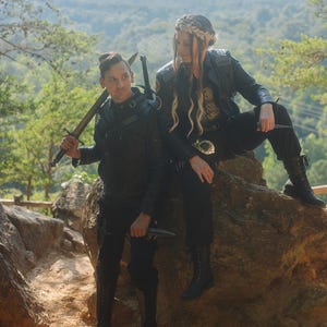 May include: Two individuals in black leather jackets and trousers, posing outdoors. One holds a sword, the other a knife. They are seated on a large rock with a forest backdrop. The image has a fantasy or cosplay theme.