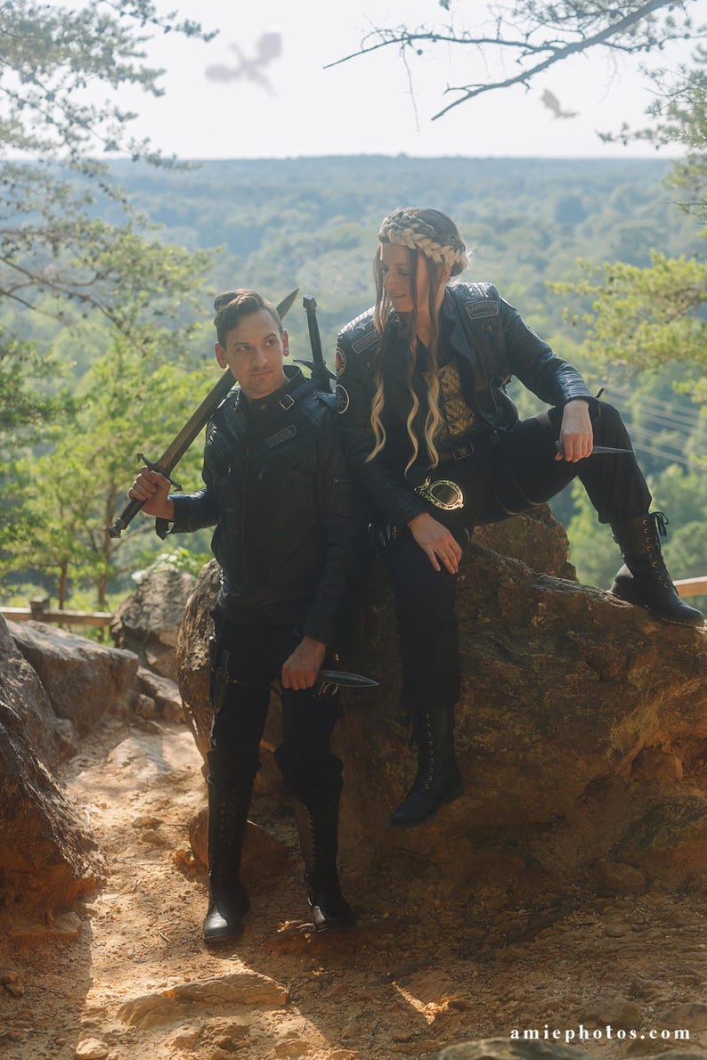 May include: Two individuals in black leather jackets and trousers, posing outdoors. One holds a sword, the other a knife. They are seated on a large rock with a forest backdrop. The image has a fantasy or cosplay theme.