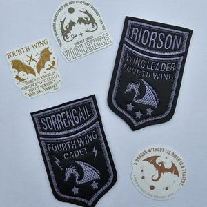 May include: A selection of embroidered patches and stickers with a fantasy theme. The patches are black with silver and grey details, featuring dragons and text like "Fourth Wing" and character names. The stickers have quotes and dragon illustrations.