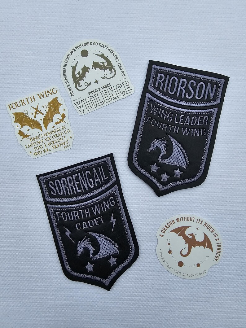May include: A selection of embroidered patches and stickers with a fantasy theme. The patches are black with silver and grey details, featuring dragons and text like "Fourth Wing" and character names. The stickers have quotes and dragon illustrations.