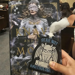 Op de afbeelding: Een boek getiteld The Knave and the Moth met een omslagillustratie van een geblinddoekte figuur met een zwaard, gecombineerd met een zwarte geborduurde patch met een gevleugelde waterspuwer en de tekst Fear Not Bartholomew met een wit lint.
