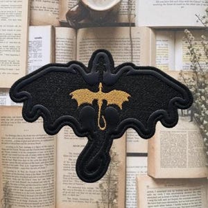 Op de afbeelding: Een zwart geborduurd embleem met een gestileerde draak met uitgespreide vleugels. Het lichaam en de vleugels van de draak zijn zwart, met een goudkleurig drakenontwerp in het midden. De patch is ontworpen om op stof te worden genaaid of gestreken.