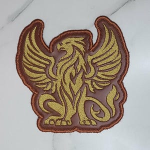 May include: A fabric patch featuring an embroidered gold griffin design on a solid brown background. The mythical creature is depicted with wings spread, a curved tail, and sharp talons. The entire shape is outlined with a thick brown stitched border.