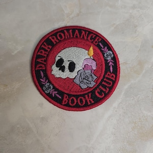 May include: A circular embroidered patch featuring a red background with a black border. The text DARK ROMANCE BOOK CLUB is stitched in red. The center displays a white skull, a lit candle with a yellow flame, and a grey rose with pink accents.