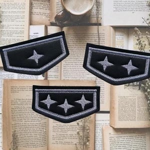 Può includere: Tre toppe nere ricamate con disegni a stella argentata. Ogni toppa ha un bordo nero e stelle argentate. Le toppe sono esposte su uno sfondo di libri aperti.