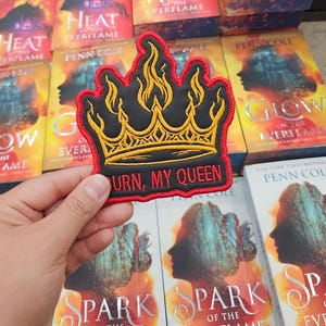 Puede incluir: Un parche bordado negro con forma de corona, detalles de llamas amarillas y un borde rojo. El texto BURN, MY QUEEN está bordado en rojo en la parte inferior. El parche se sostiene frente a un fondo de varios ejemplares de los libros Heat of the Everflame, Glow of the Everflame y Spark of the Everflame.