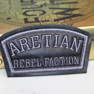 May include: A black faux leather patch with a curved top edge and a rectangular base. The patch features embroidered light gray text that reads ARETIAN in an arched font and REBEL FACTION in a straight line below, bordered by gray stitching.