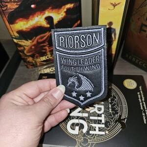 May include: A black and white patch featuring the text "RIORSON WING LEADER FOURTH WING" and a dragon design. The patch is held in front of a book titled "Fourth Wing" and other books.