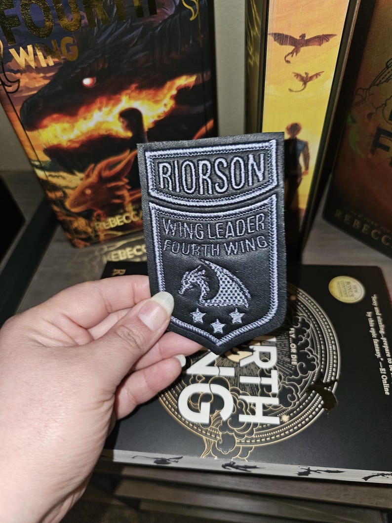 May include: A black and white patch featuring the text "RIORSON WING LEADER FOURTH WING" and a dragon design. The patch is held in front of a book titled "Fourth Wing" and other books.