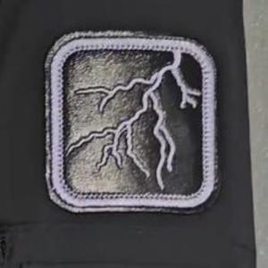 May include: A square-shaped patch featuring a white embroidered lightning bolt design on a black background. The patch has a thick white border with visible stitching and is attached to a solid black fabric surface.