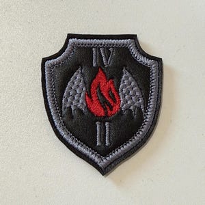 May include: A shield-shaped black patch with a grey embroidered border. The center features a red flame graphic flanked by two grey textured wings. The Roman numeral IV is embroidered above the flame, and the Roman numeral II is below the flame.