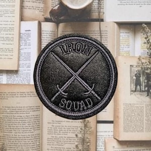 May include: A black circular patch with the words "IRON SQUAD" and two crossed swords embroidered in white. The patch is set against a backdrop of open vintage books and dried flowers.
