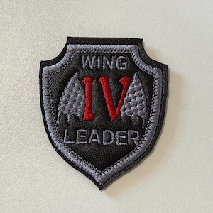 Puede incluir: Un parche bordado en forma de escudo con una base negra de aspecto cuero. Presenta el texto WING en gris, el número romano IV en rojo y LEADER en gris. El borde está cosido con hilo gris y dos iconos de alas grises flanquean el IV rojo.