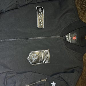 May include: Black bomber jacket with multiple patches and star accents. The jacket features the name "RIORSON" on several patches, along with other text and a shield design. A zipper runs down the front.