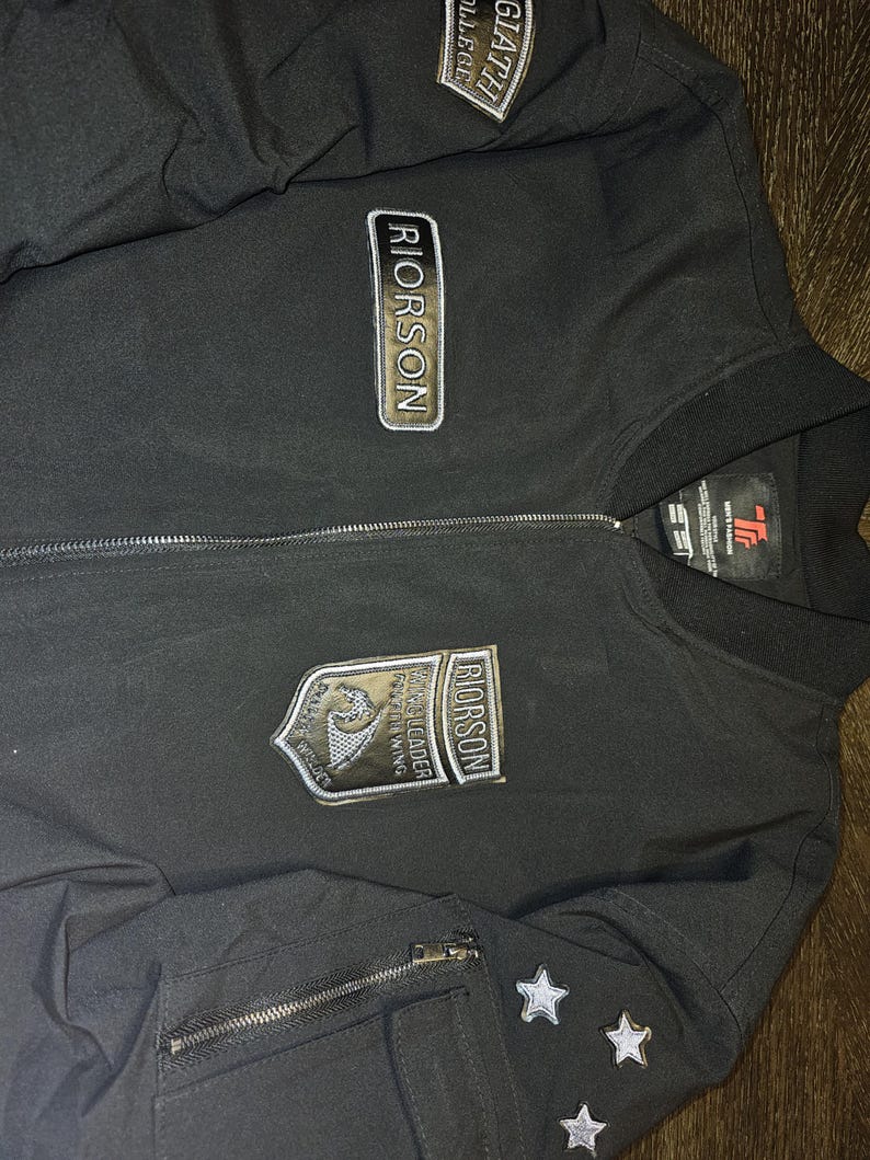 May include: Black bomber jacket with multiple patches and star accents. The jacket features the name "RIORSON" on several patches, along with other text and a shield design. A zipper runs down the front.