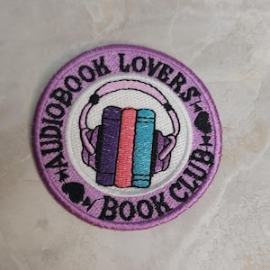 May include: A circular embroidered patch with a purple border and white center. It features a graphic of three vertical books in purple, pink, and teal, topped with light pink headphones. The text reads AUDIOBOOK LOVERS BOOK CLUB in black.