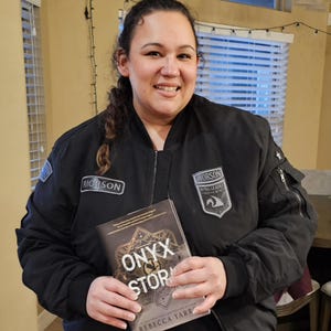 May include: A person wearing a black bomber jacket with patches, holding a book titled "Onyx Storm" by Rebecca Yarra. The jacket has the word "Rioroson" on it. The book cover features a geometric design.