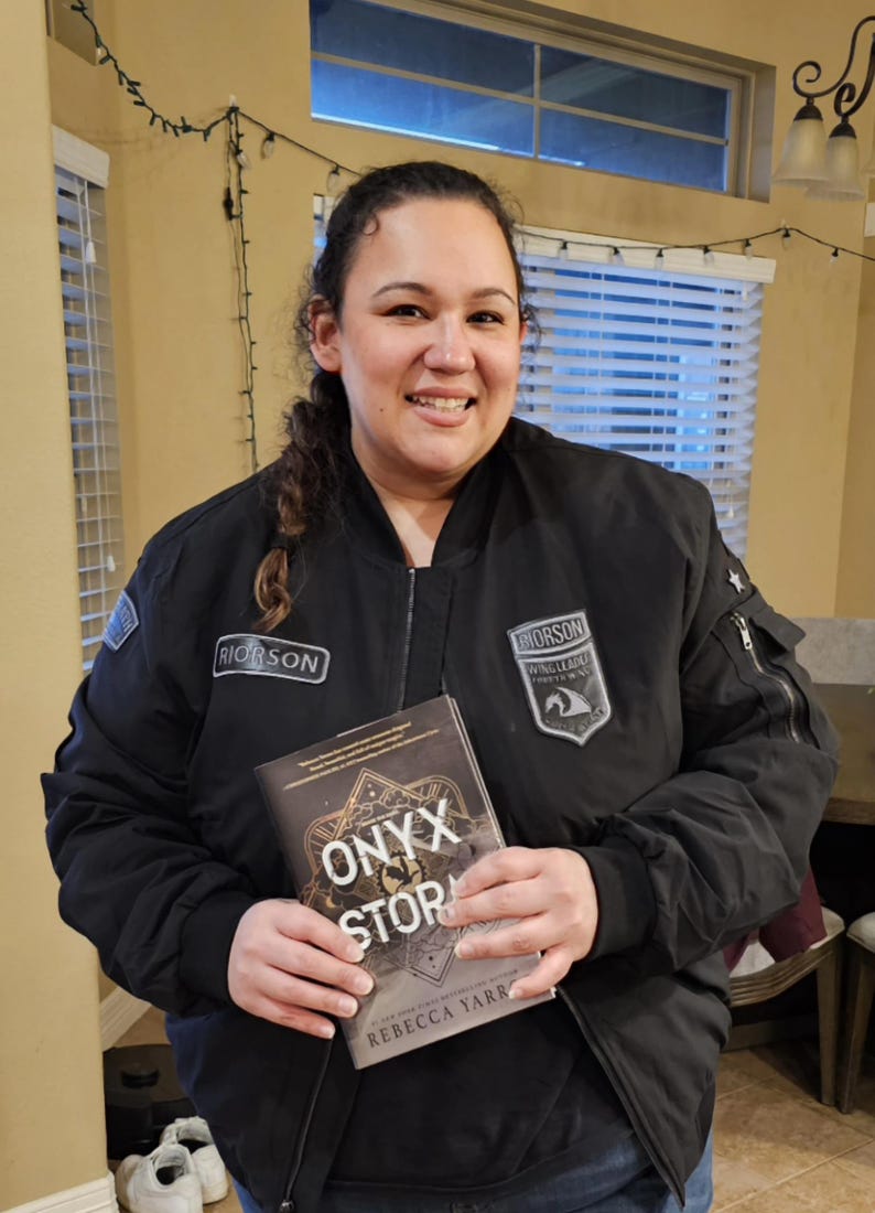 May include: A person wearing a black bomber jacket with patches, holding a book titled "Onyx Storm" by Rebecca Yarra. The jacket has the word "Rioroson" on it. The book cover features a geometric design.
