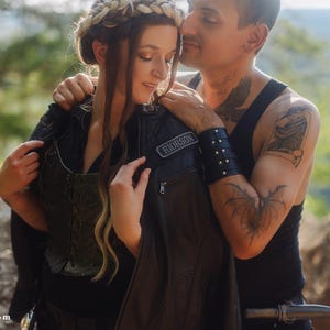 May include: A couple embracing outdoors. The woman wears a black leather jacket with a patch that reads "RIORSON". The man has tattoos on his arm and wears a black vest. The woman has a braided hair crown.
