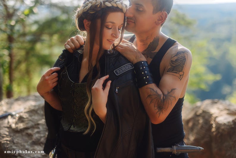May include: A couple embracing outdoors. The woman wears a black leather jacket with a patch that reads "RIORSON". The man has tattoos on his arm and wears a black vest. The woman has a braided hair crown.