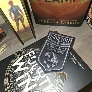 May include: A black and silver patch with the text "RIORSON WING LEADER FOURTH WING" and a dragon emblem. The patch is on a book with gold lettering and a black cover. A book with the title "IRON FLAME" is also visible.