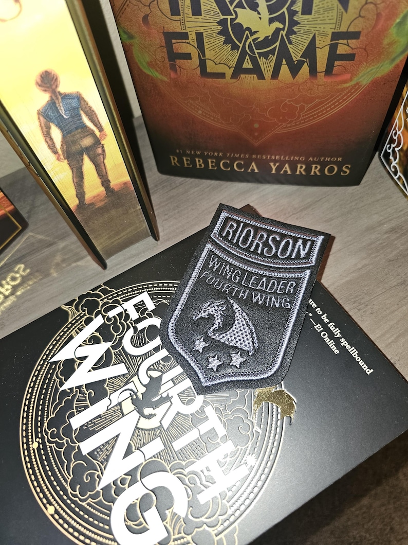 May include: A black and silver patch with the text "RIORSON WING LEADER FOURTH WING" and a dragon emblem. The patch is on a book with gold lettering and a black cover. A book with the title "IRON FLAME" is also visible.