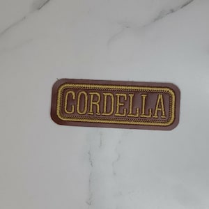 May include: A rectangular patch with rounded corners featuring a dark brown background and a gold embroidered border. The name CORDELLA is stitched in gold capital letters in the center of the patch, creating a clear and legible design.