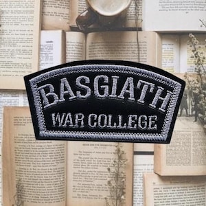 Könnte beinhalten: Schwarzer Aufnäher mit weiß gesticktem Text „BASGIATH WAR COLLEGE“. Der Aufnäher hat eine gebogene Oberseite und eine rechteckige Form. Der Hintergrund besteht aus offenen Büchern.