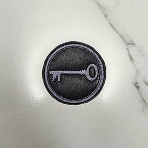 May include: A circular embroidered patch featuring a black leather-like center with a light purple key icon stitched in the middle. The patch is bordered by a thick, light purple stitched edge, creating a distinct contrast against the dark center.