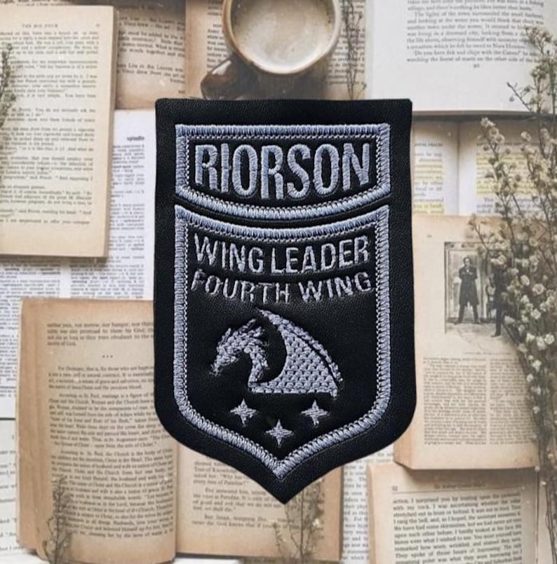 May include: A black and grey embroidered patch with the words "RIORSON WING LEADER FOURTH WING". The patch has a dragon design and three stars. The shape is a shield.